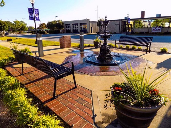 Street-level view of a decorative water fountain and seating area in downtown Angier, North Carolina with benches, brickwork, and landscaped planters.
