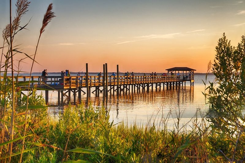 People fishing on a wooden pier at sunset over calm coastal waters in Wilmington, NC.